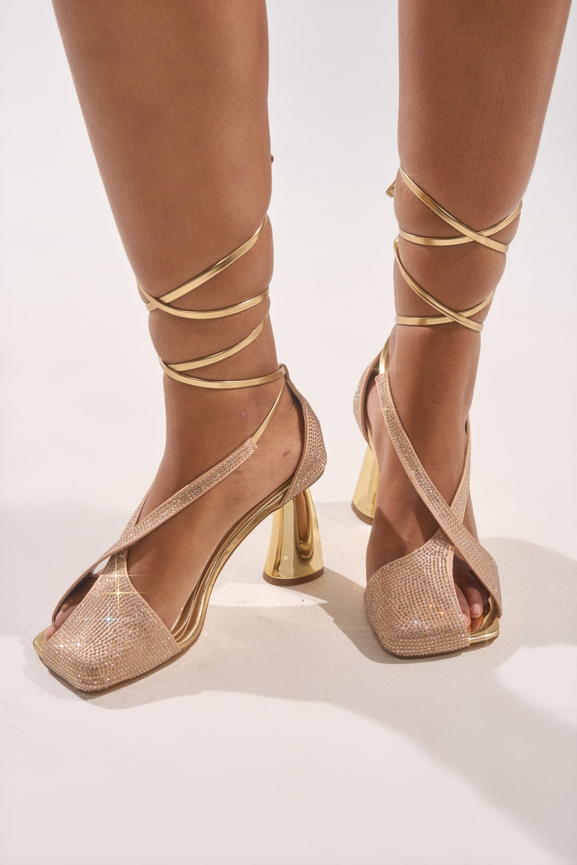 A pair of feet wearing the AZALEA WANG BUILANY GOLD RHINESTONE CHUNKY PUMP with sparkly criss-cross straps and thick metallic gold heels stands on a light-colored surface.