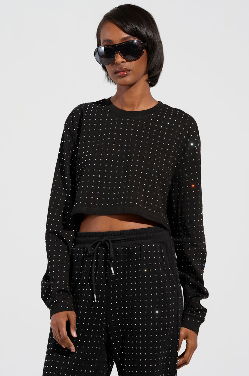 A person sporting the Buttercup Long Sleeve Rhinestone Embellished T-Shirt in black, featuring large sunglasses, exudes elegance in a sparkling outfit. The attire includes a rhinestone-embellished cropped silhouette with matching pants adorned with small sequins, set against a plain light gray background.