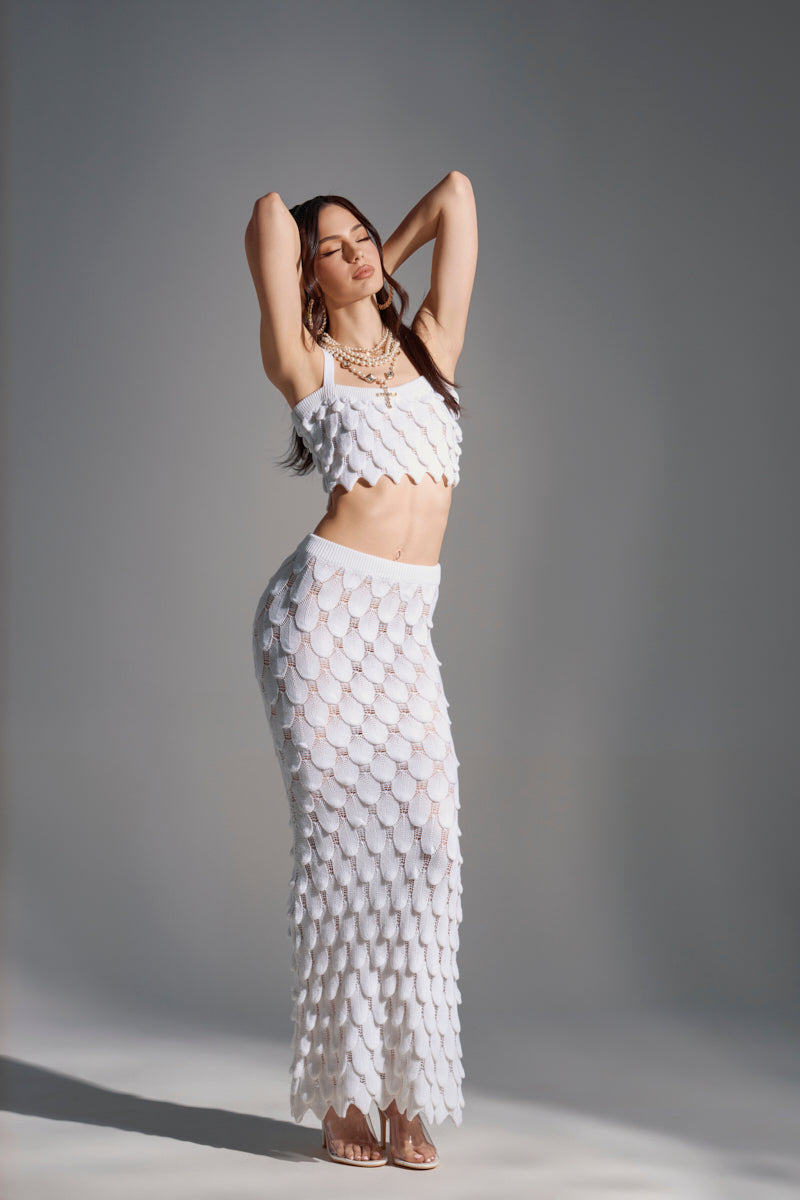 A woman poses in a studio against a gray background, wearing the A MOMENT APART MAXI SKIRT with a matching white scallop-patterned crochet knit crop top, layered necklaces, and clear high-heeled sandals, hands raised behind her head.