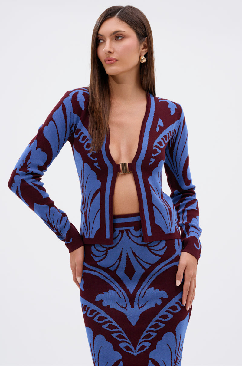 A woman models the GIVE ME LOVE SWEATER, featuring a deep V-neck with a gold clasp and a matching high-waisted skirt in bold blue and burgundy leaf damask, posing against a white background with her long brown hair down. 0