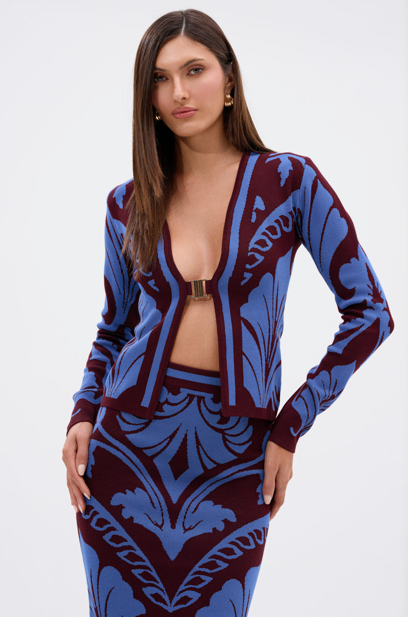 A woman with straight brown hair wears the GIVE ME LOVE SWEATER in rich burgundy with a gold clasp, paired with a matching high-waisted damask skirt, standing against a plain light background. 1