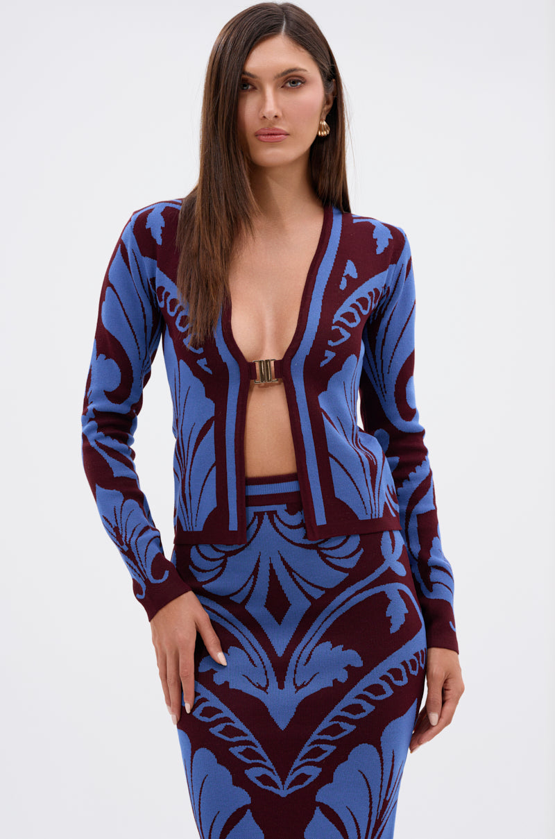 A woman with straight brown hair wears the GIVE ME LOVE SWEATER, a blue and burgundy patterned cardigan with a deep neckline and center clasp, paired with a matching skirt. She stands against a plain white background. 2