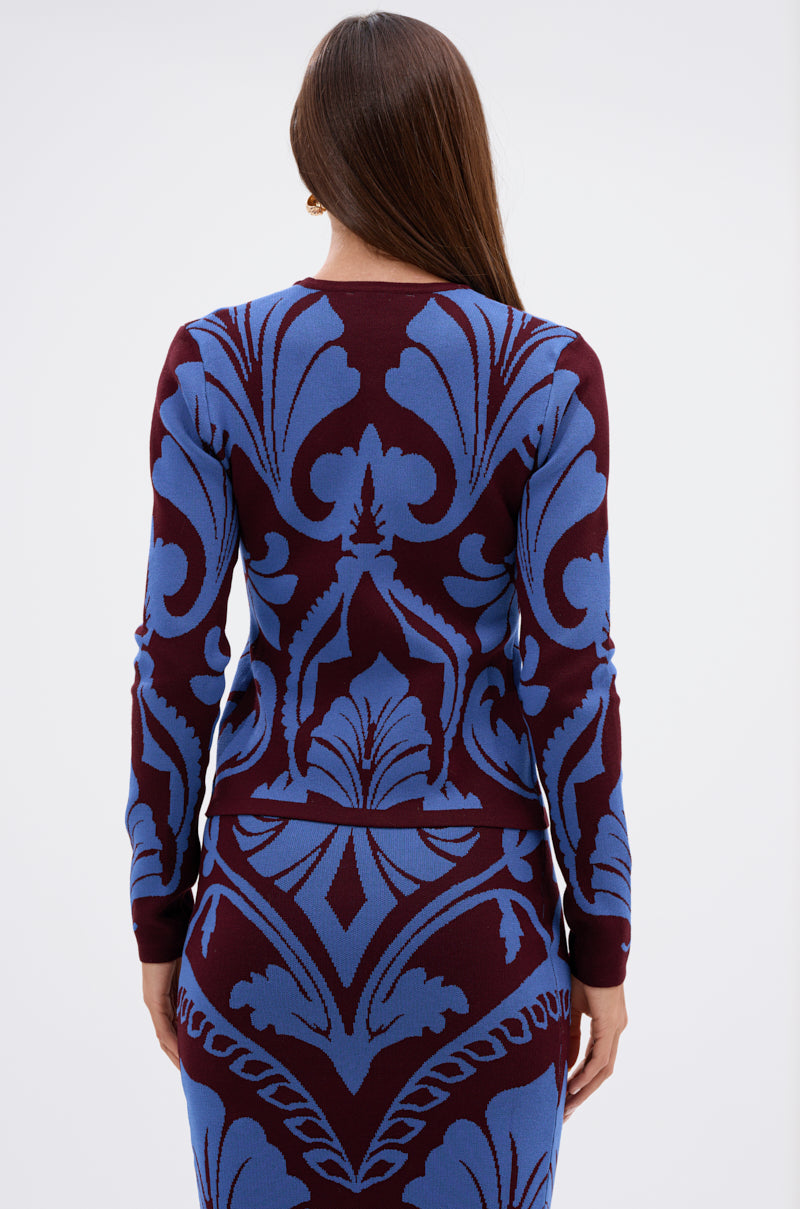 A woman with long brown hair is seen from behind, wearing the GIVE ME LOVE SWEATER, a fitted long-sleeve piece with bold blue and burgundy damask patterns against a plain white background. 4