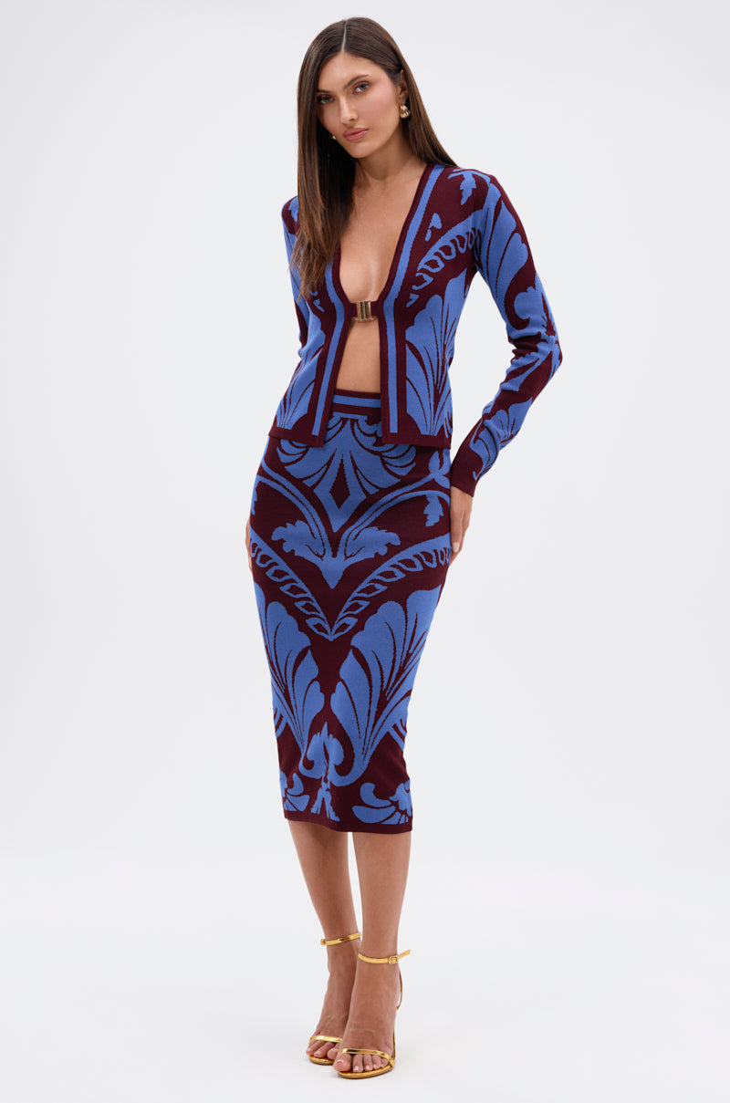 A woman wearing a form-fitting, deep V-neck GIVE ME LOVE SWEATER with a matching midi skirt in ornate blue and burgundy damask stands in gold strappy heels, one arm at her side and the other bent. 7