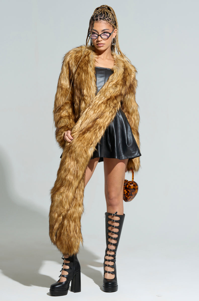 A person confidently stands wearing a long, LANGON High Low Bolero by AKIRA Label over a black leather dress and tall lace-up platform boots. They also sport stylish glasses and carry a heart-shaped bag.