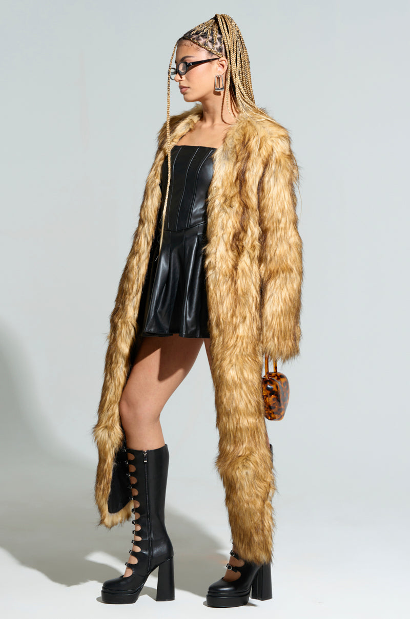 A fashionably dressed woman in an AKIRA LANGON High Low Bolero paired with a black leather dress, and knee-high lace-up boots. She sports braided hair and glasses while carrying a small purse. The background is a plain light color.