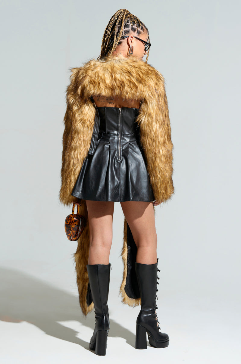 A woman stands with her back to the camera, showcasing a brown LANGON HIGH LOW BOLERO over a black leather mini dress. Her knee-high black boots and sunglasses enhance her stylish ensemble, completed with an AKIRA Label Langon bag. Her braided hair adds flair to the high-low design of her look.