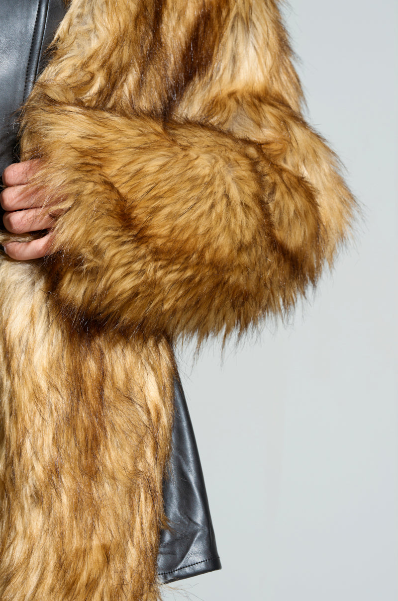 A close-up of a person wearing the LANGON HIGH LOW BOLERO, featuring thick, fluffy faux fur and a sleek black leather panel. The person's hand is partially visible, holding the edge of the bolero. The background is a plain, light gray, emphasizing the high-low design of this chic outerwear.