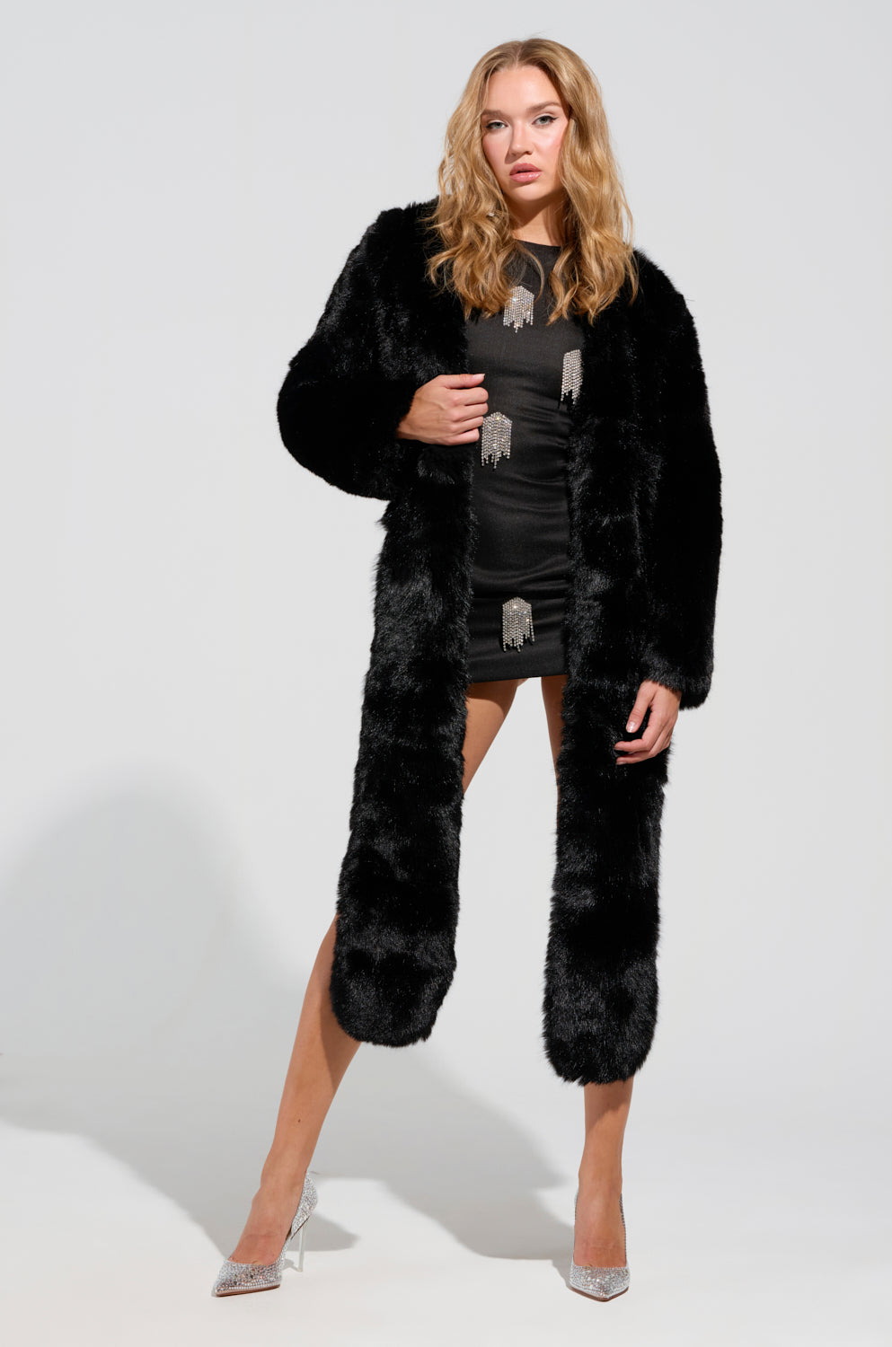 Dressed in the stylish Business In The Front Asymmetrical Fur Bolero, a woman exudes confidence with her sophisticated look, sporting a short dress adorned with tassels. She strikes a poised stance with one leg forward, wearing silver high heels against a simple white background—a flawless ensemble for the holiday season.