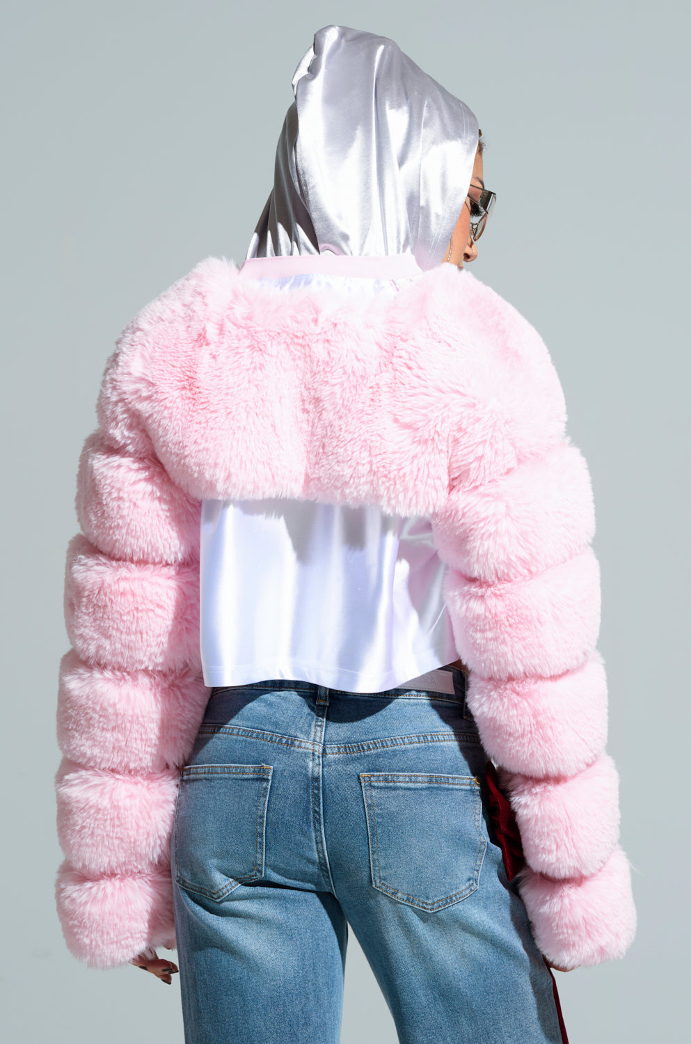 A person wearing the LADIES NIGHT FAUX FUR BOLERO in vibrant pink poses against a plain background. They complement the look with a shiny silver headscarf and blue jeans featuring a side stripe. The image, captured from behind, emphasizes the jacket's texture, ideal for a stylish ladies' night out.