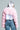 A person wearing the LADIES NIGHT FAUX FUR BOLERO in vibrant pink poses against a plain background. They complement the look with a shiny silver headscarf and blue jeans featuring a side stripe. The image, captured from behind, emphasizes the jacket's texture, ideal for a stylish ladies' night out.