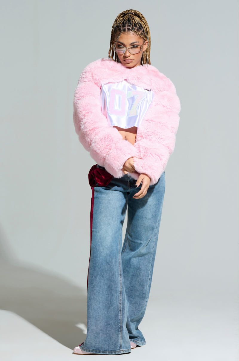 With braided hair styled confidently, the person is dressed in the LADIES NIGHT FAUX FUR BOLERO—a velvet smooth pink fluffy jacket—paired with a white crop top featuring text and wide-leg jeans. Completing the chic look are stylish sunglasses and vibrant high heels, all posed against a light gray backdrop. Ideal for exuding elegance and flair on ladies' night.