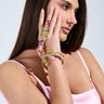 A woman with long brown hair in a pink top rests her hand against her face, revealing the MY SECRET GARDEN LUXE GEM BRACELET RING COMBO, a colorful piece with textured floral designs. The light background highlights her style.