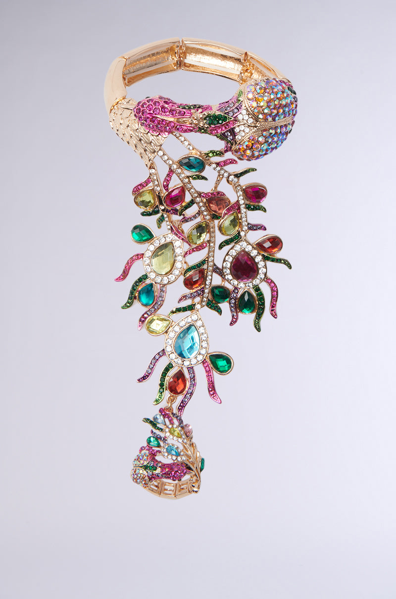 Introducing the Wild Nights Bracelet with Ring, a statement piece featuring rhinestone-encrusted peacock motifs. This ornate design showcases vibrant pink, green, and blue gemstones against a light gray background, capturing a peacock's head and flowing feathers with intricate detail. 2