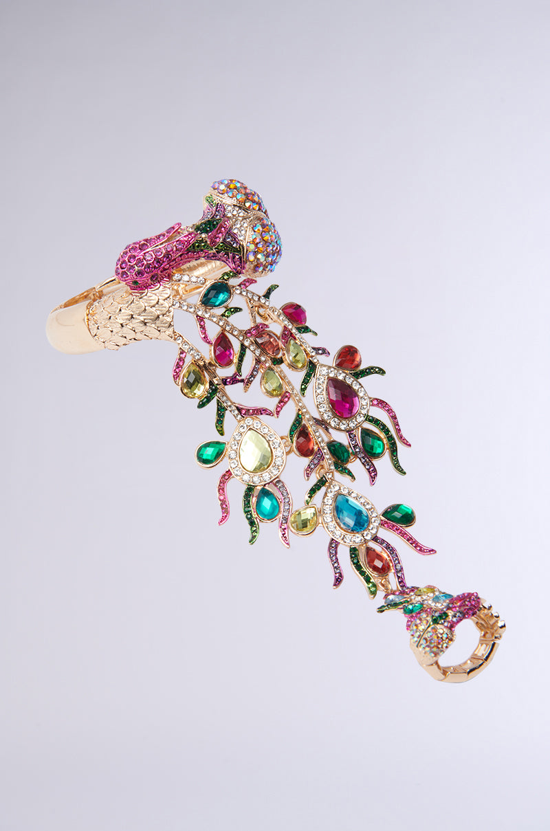 Discover the Wild Nights Bracelet with Ring, a statement piece showcasing a rhinestone-encrusted peacock adorned with multicolored gemstones. Its beautiful tail cascades in green, pink, and blue stones set against a light gray backdrop for an extra touch of elegance. 3