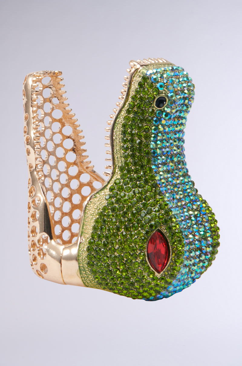 The BITE ME CUFF BRACELET features an intricately designed gold structure shaped like an open crocodile's head, embellished with multicolor rhinestones. A red gem is placed as the eye, creating a striking contrast against the metallic and jeweled textures. 0