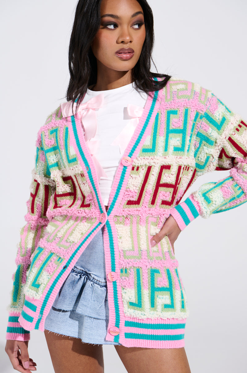 Wearing the FREE SPIRIT CARDIGAN with an oversized silhouette and pastel teal, pink, and white geometric patterns, she pairs it with a light pink top and denim shorts. Her long dark hair cascades as she turns against a plain backdrop.