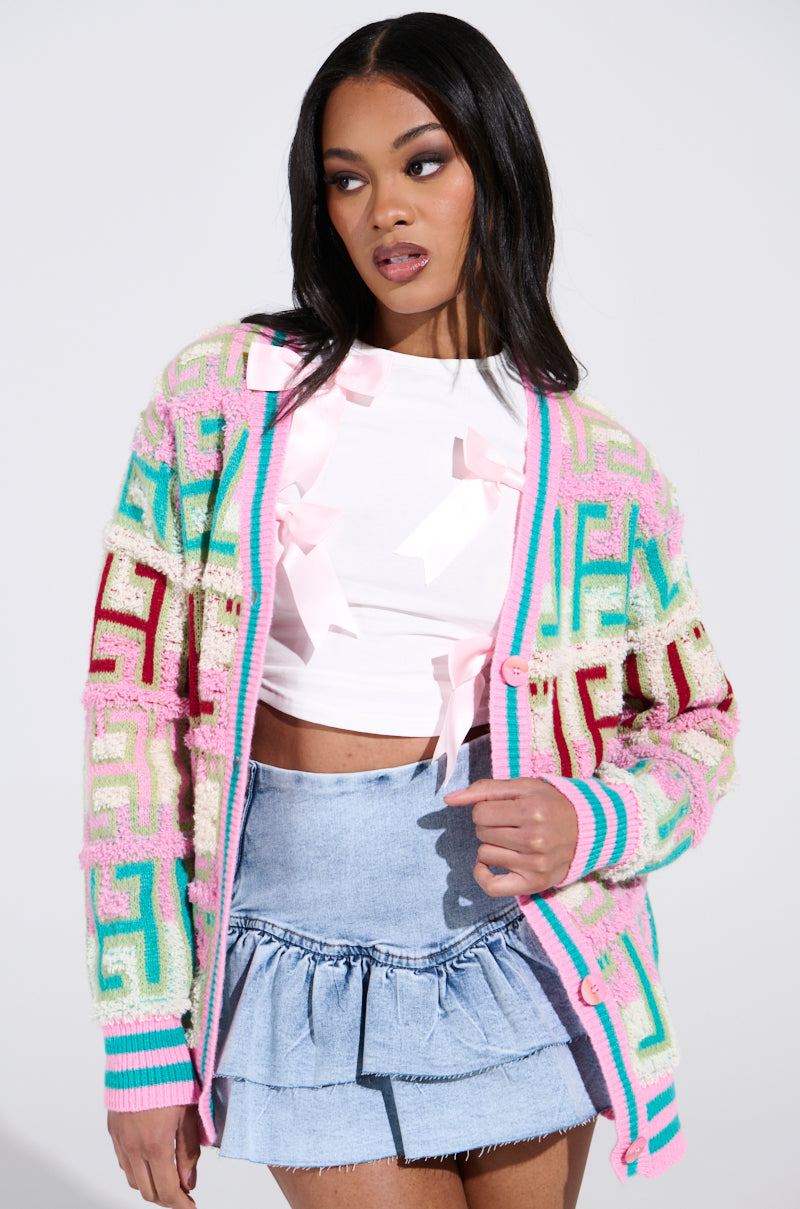 A person with long dark hair wears the FREE SPIRIT CARDIGAN, known for its colorful patterns and oversized silhouette, paired with a white top featuring bows and a denim skirt. They look to the side against a plain background.