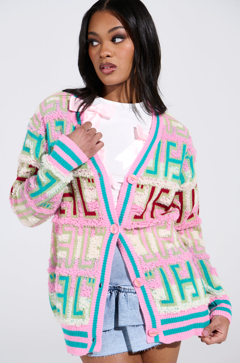A woman models the FREE SPIRIT CARDIGAN, showcasing its oversized color block design in pink, blue, and beige with geometric patterns. It features pink buttons and is worn over a white top and denim shorts. She poses with one hand on the collar, gazing to the side.