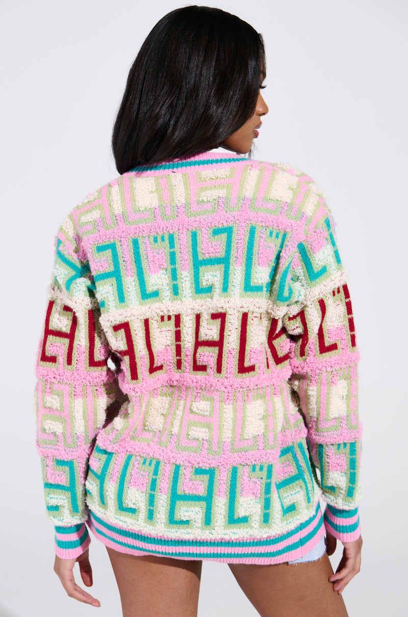 A person with long black hair faces away from the camera, wearing an oversized FREE SPIRIT CARDIGAN adorned with colorful geometric patterns in pink, green, blue, and red. The cardigan's hem has a striped design contrasting a plain white background.