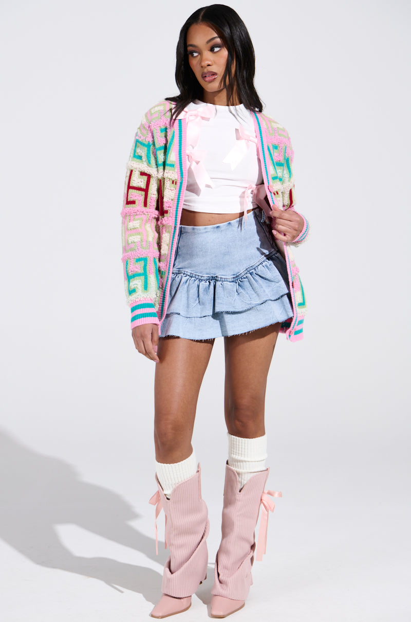 A person confidently wears a FREE SPIRIT CARDIGAN with a colorful color block design, paired with a white top featuring pink ribbon details, a light blue denim skirt, and pink high-heeled boots adorned with bows, all against a plain white background.
