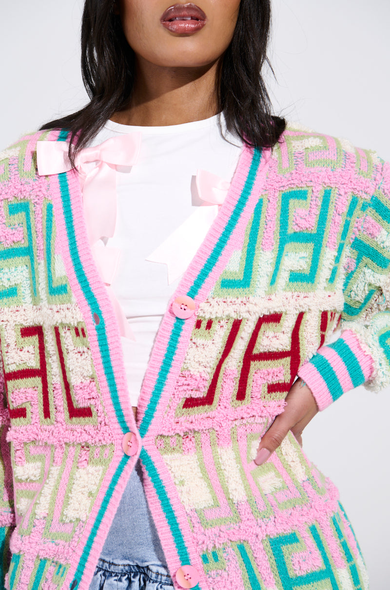 A person dons the vibrant FREE SPIRIT CARDIGAN, featuring an oversized silhouette and a geometric color block in pink, turquoise, and cream over a white shirt. The cardigan's pink buttons and the shirt's pink ribbon detail perfectly complement their long, dark hair.