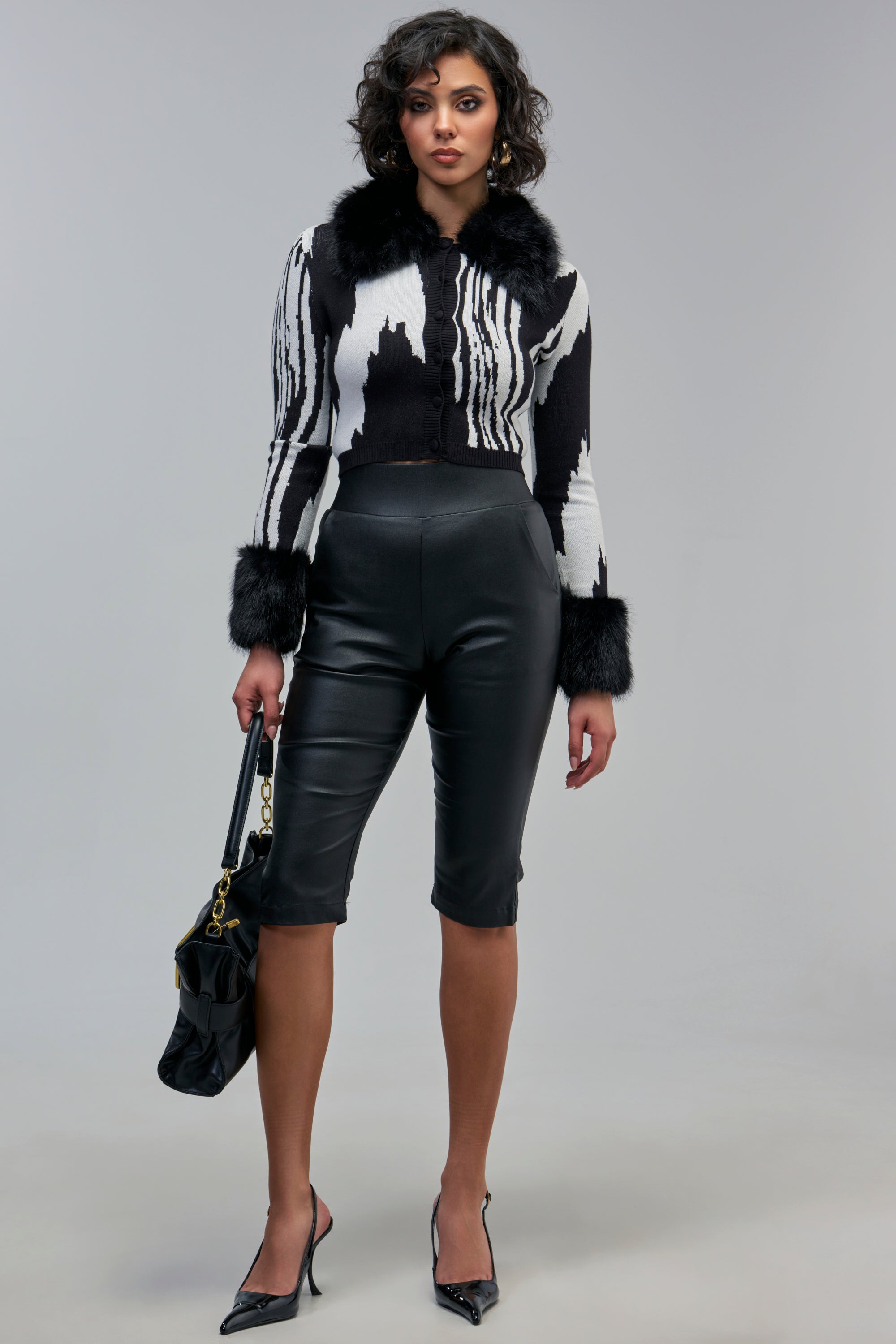 A woman wearing a black-and-white patterned cardigan with faux fur trim, AMMO X AKIRA BIG BOOTY PLEATHER CAPRI WITH 4 WAY STRETCH, and black heeled pumps, holding a black handbag.