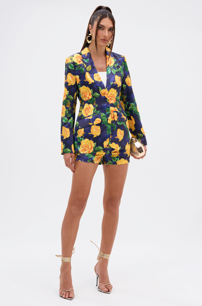 A woman poses against a white background in a navy blazer, DOZEN ROSES SATIN SHORTS IN NAVY with a bold yellow rose print, gold hoop earrings, a gold clutch, and strappy gold high-heeled sandals. 5