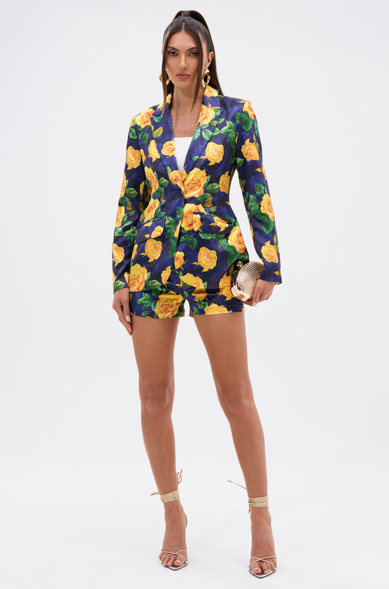 A woman with straight hair and a confident look models the DOZEN ROSES SATIN SHORTS IN NAVY—featuring a bold yellow rose print—styled with a navy blazer, gold heels, and a matching gold clutch against a white background. 6
