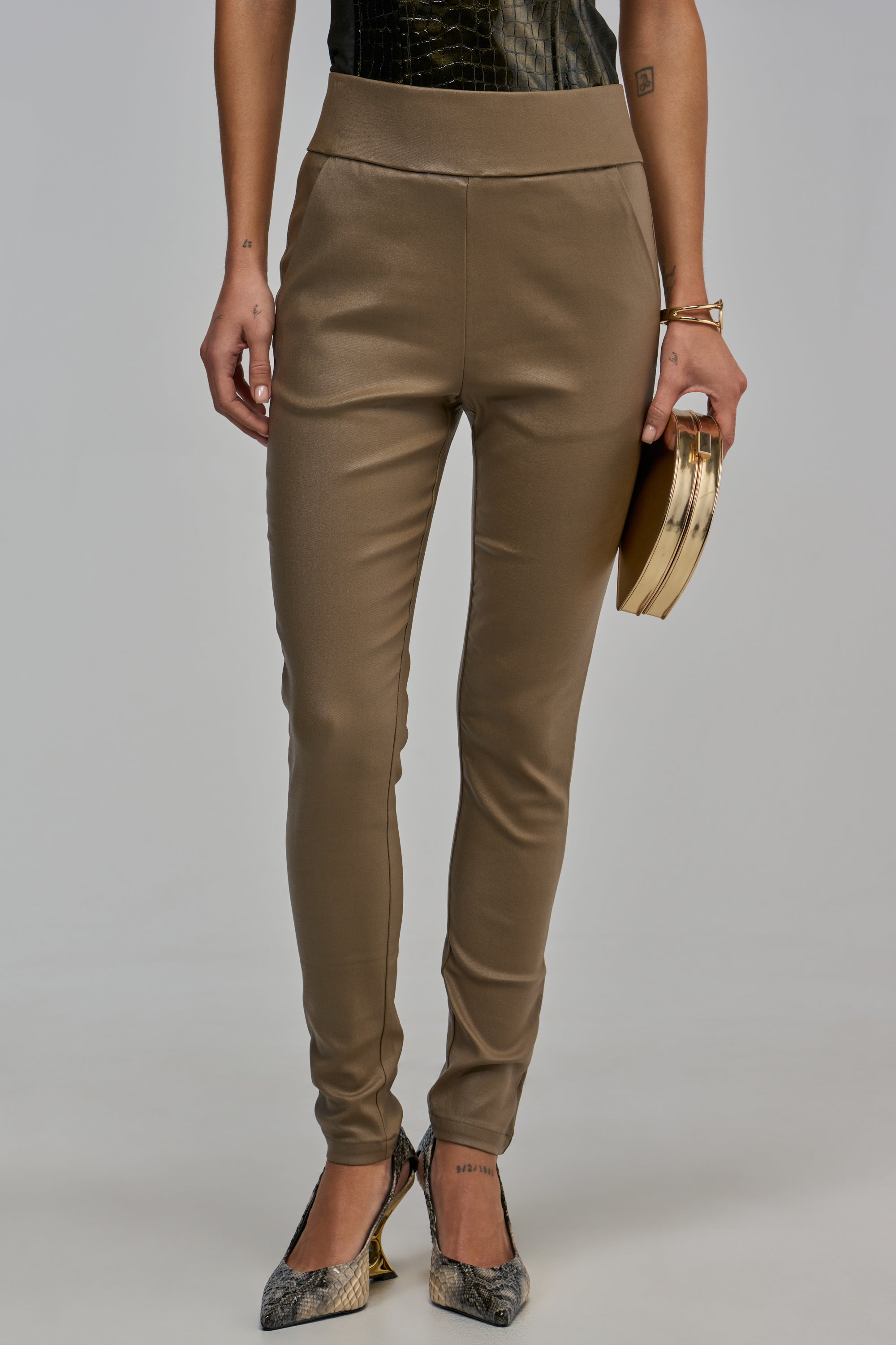 AMMO X AKIRA BIG BOOTY HIGH WAIST PLEATHER PANT IN OLIVE in a skinny-leg silhouette, worn with a black top, a gold clutch, and snakeskin print heels.