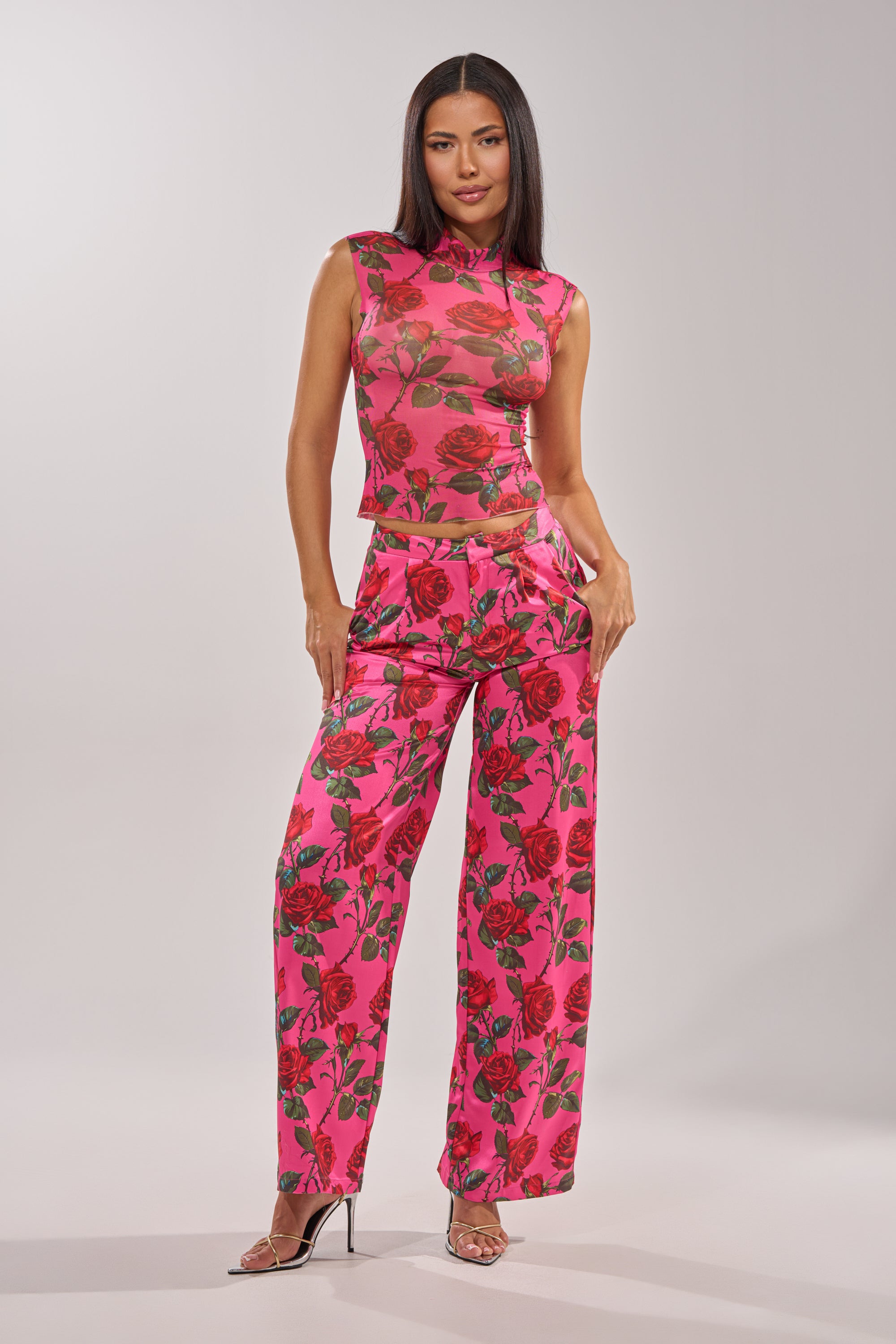A woman models the DOZEN ROSES SATIN PANT IN FUCHSIA, a high-waisted wide-leg pant with a red rose print, paired with a matching sleeveless top. She completes the look with clear heels and wears her hair down against a plain background. 12