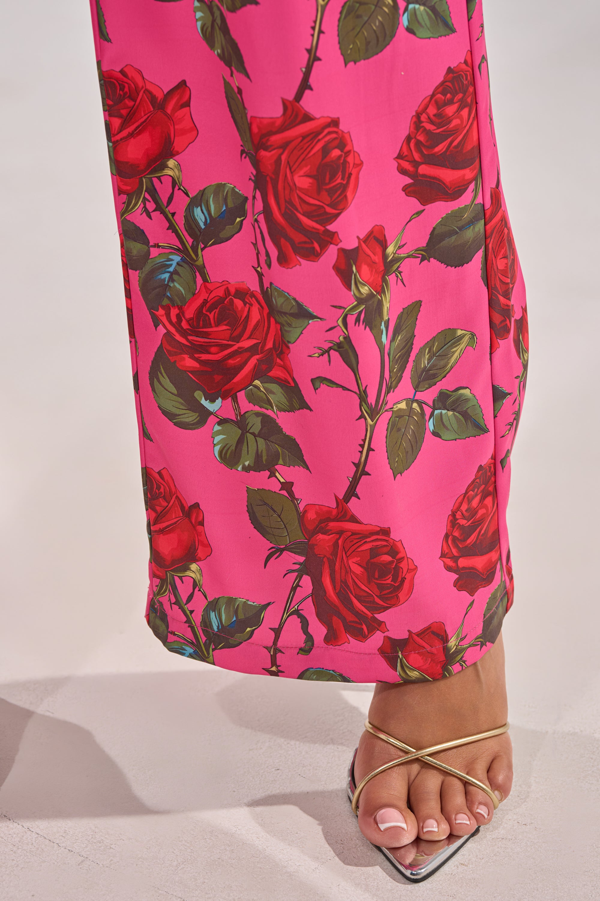 A person wearing DOZEN ROSES SATIN PANT IN FUCHSIA and silver strappy high-heeled sandals stands on a light-colored floor, with only their lower leg and foot visible. 14