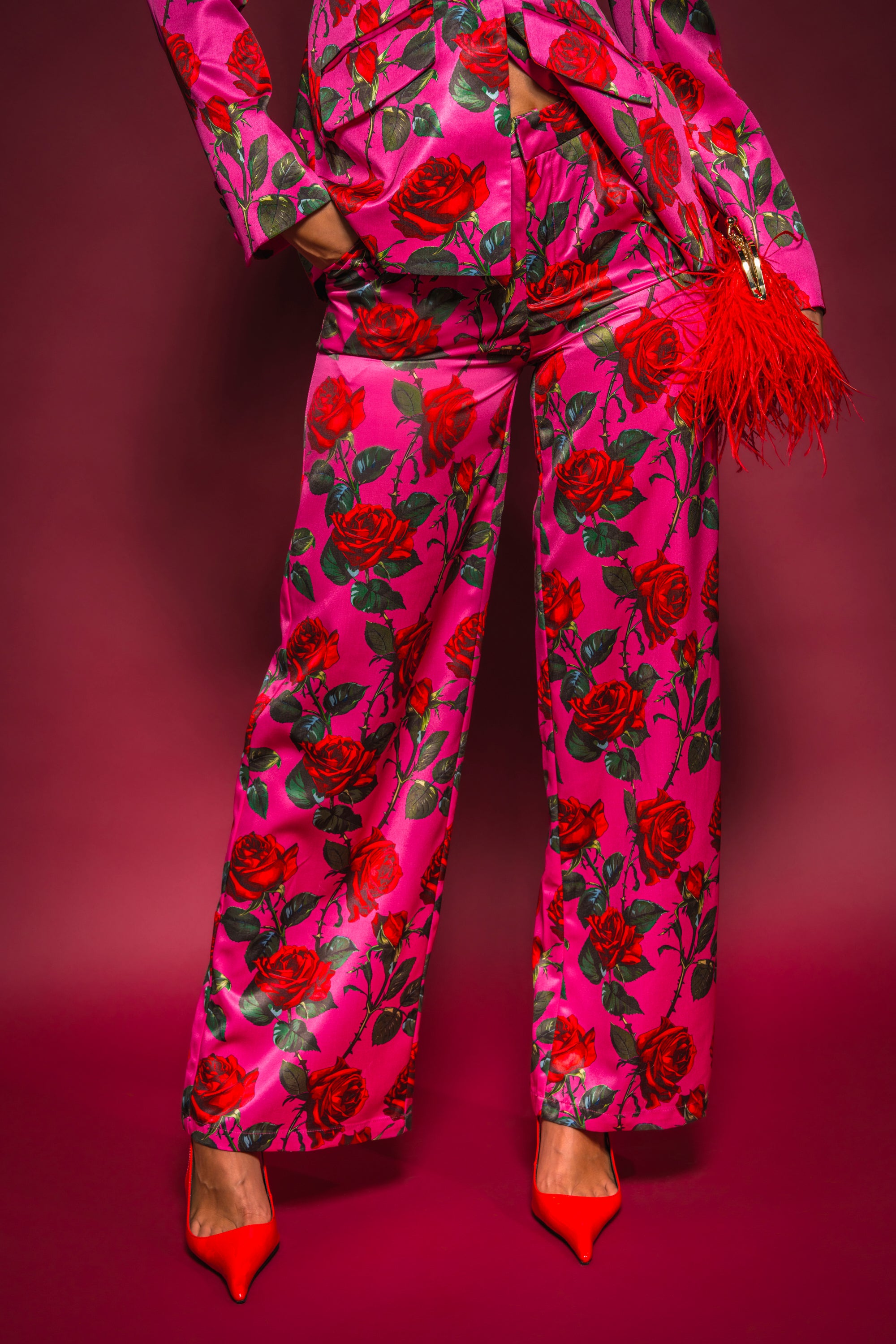 A person wears the DOZEN ROSES SATIN PANT IN FUCHSIA, styled with red pointed-toe heels and a red feathered clutch, posing against a pink-red backdrop. 1