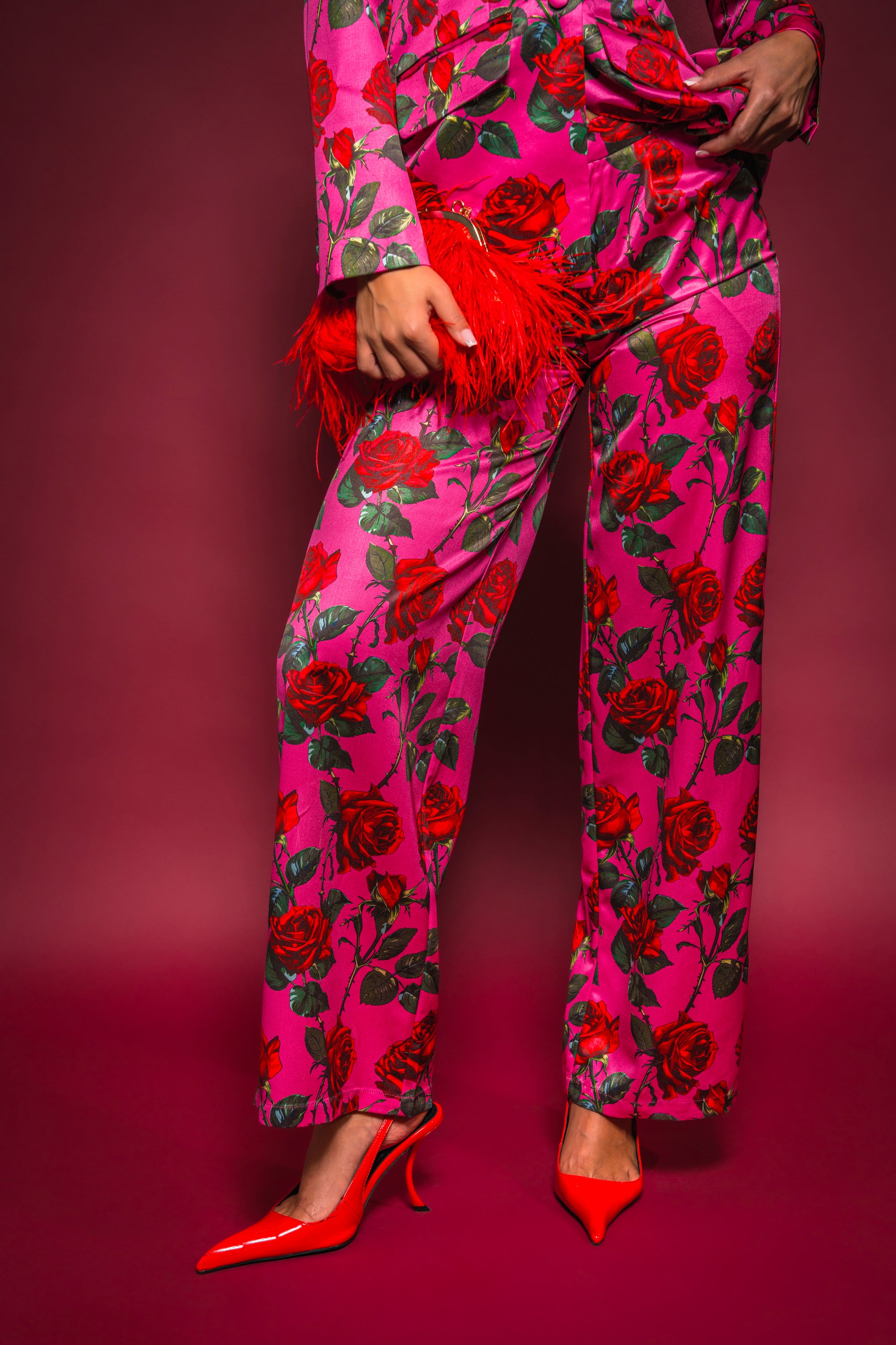 A person poses against a red background, wearing the DOZEN ROSES SATIN PANT IN FUCHSIA with rose prints and red high heels, holding a red feathered clutch. Only the lower half of the body is shown. 2