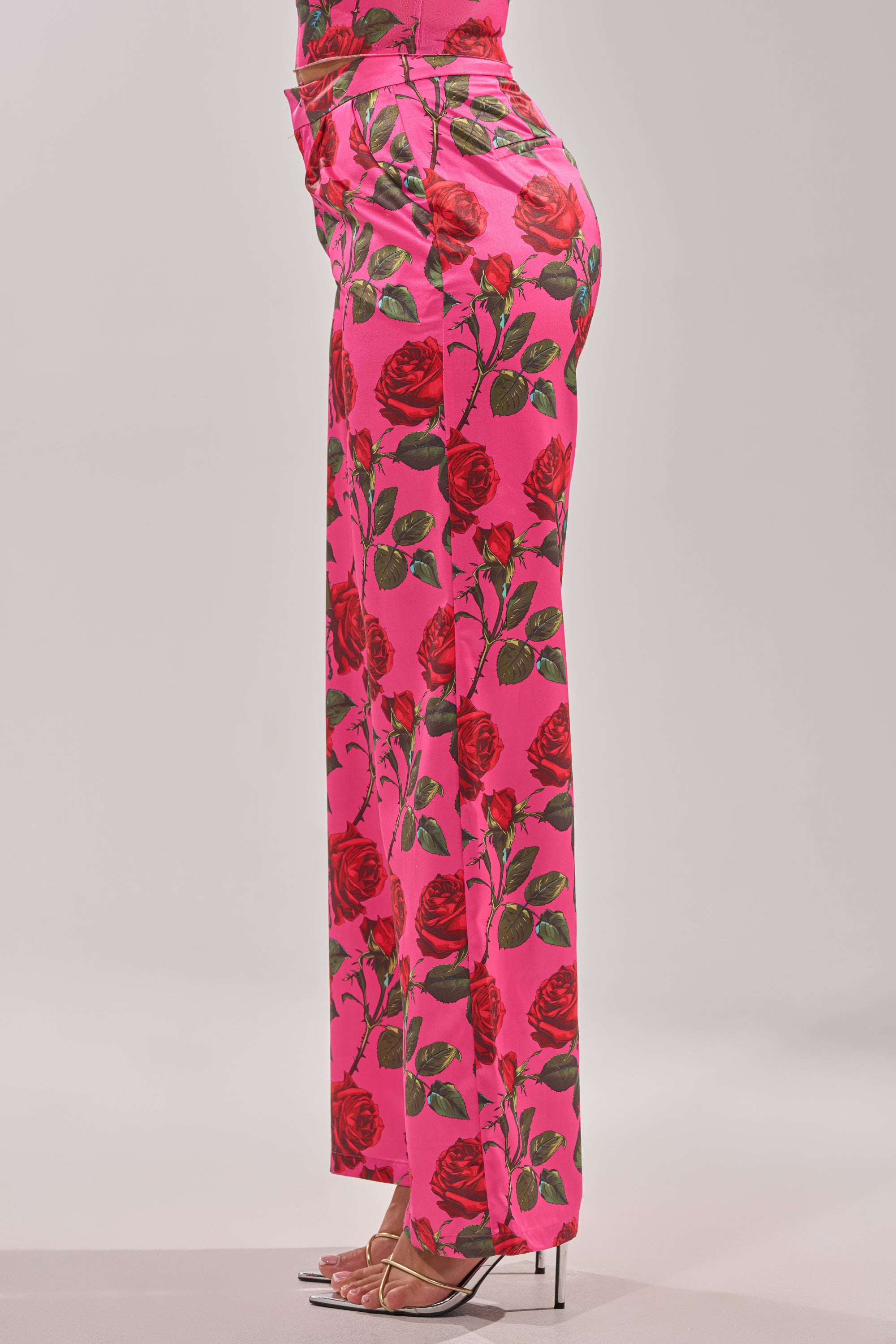 A person stands in profile wearing the DOZEN ROSES SATIN PANT IN FUCHSIA, featuring a wide-leg silhouette and rose-print pattern, paired with strappy clear high heels against a plain background. 7