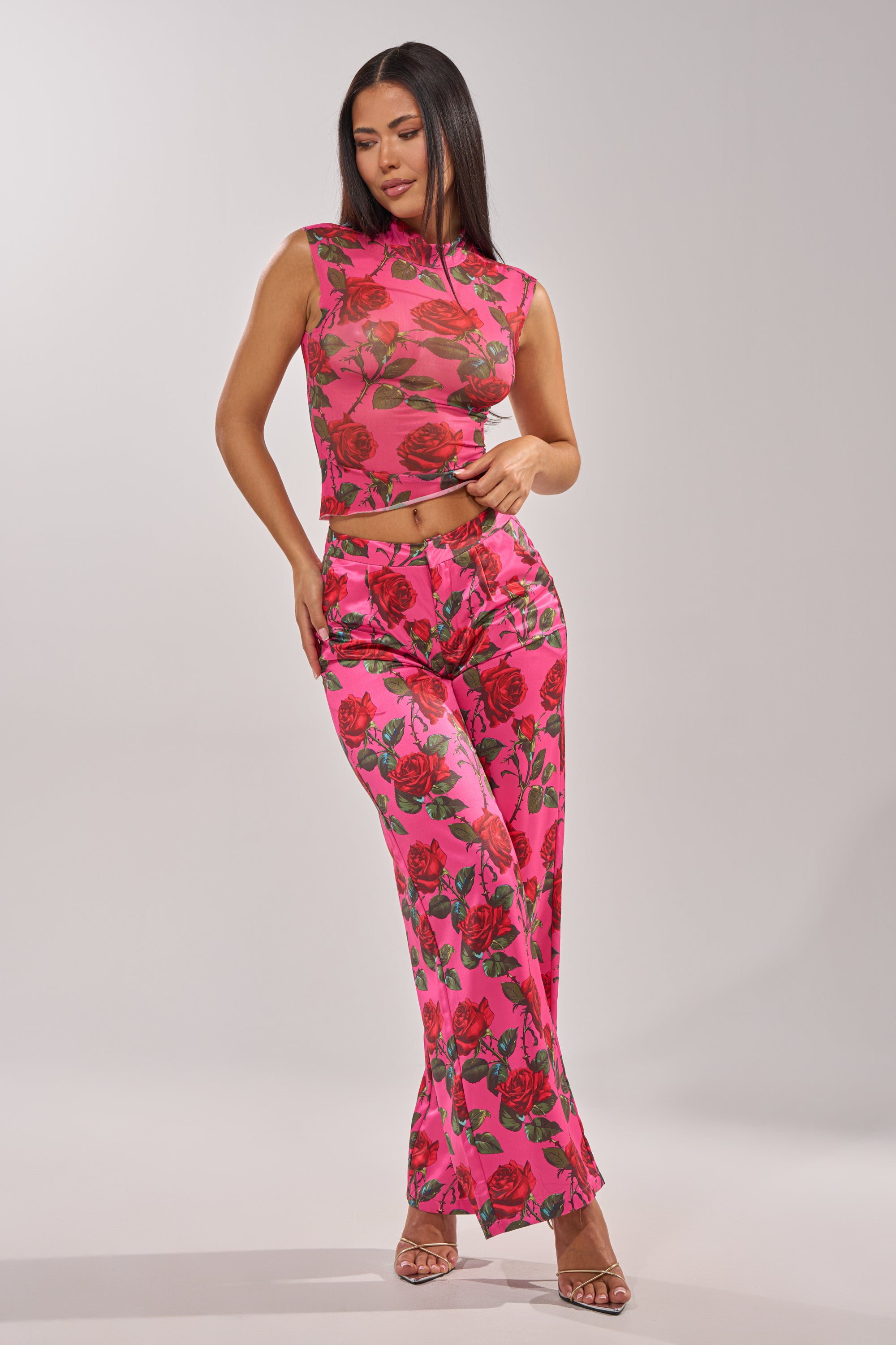 A woman models the DOZEN ROSES SATIN PANT IN FUCHSIA, a high-waisted wide-leg style with a rose print, paired with a fitted pink crop top and open-toed heels against a plain light background. 11