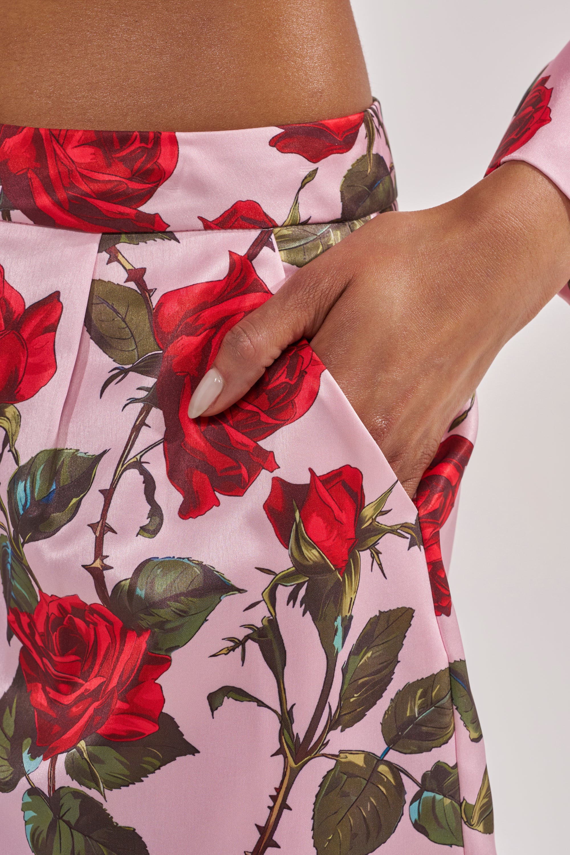 A hand with manicured nails in a pocket of the DOZEN ROSES SATIN SHORTS IN LIGHT PINK, featuring a bold red rose print. The model has medium brown skin and the softly lit background is plain. 7