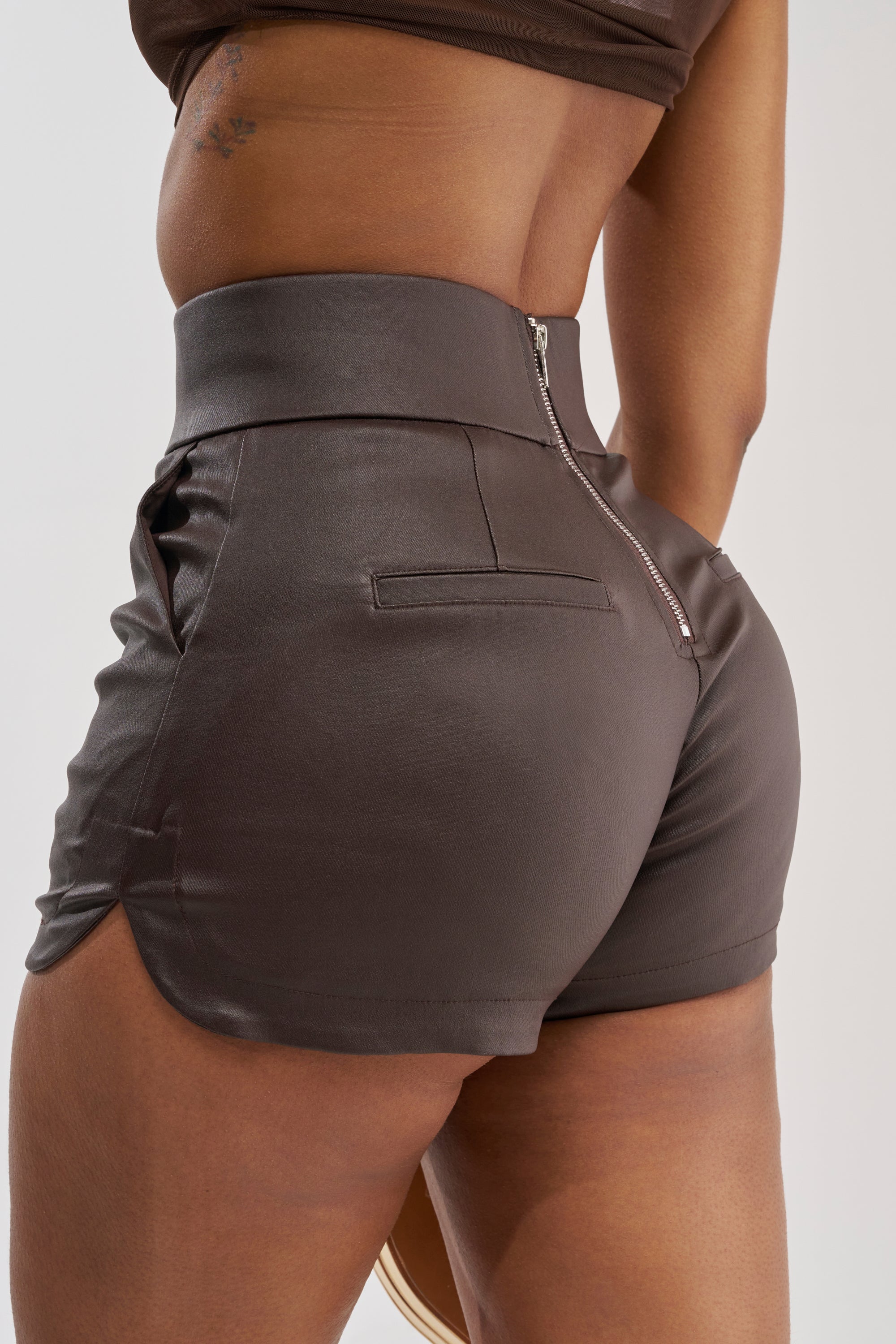 A person wearing the AMMO X AKIRA BIG BOOTY PLEATHER SHORTS WITH 4 WAY STRETCH—high-waisted, fitted, dark brown faux leather shorts featuring a visible back zipper and side pockets—stands with their hand in a pocket.