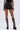 Wearing AMMO X AKIRA BIG BOOTY PLEATHER SHORTS WITH 4 WAY STRETCH in black, a black crop top, and knee-high platform boots with silver buckles, a person stands against a plain light background.