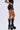 A person wears AMMO X AKIRA BIG BOOTY PLEATHER SHORTS WITH 4 WAY STRETCH, a black sleeveless top, and tall black lace-up platform boots with side zippers, standing against a plain light background.