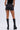 A person faces away in AMMO X AKIRA BIG BOOTY PLEATHER SHORTS WITH 4 WAY STRETCH, paired with a black top and tall black platform boots featuring laces and side zippers, set against a plain light background.