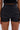A person wears AMMO X AKIRA BIG BOOTY PLEATHER SHORTS WITH 4 WAY STRETCH in high-rise black pleather, paired with a black cropped top, shown from mid-torso to mid-thigh against a neutral background.