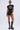 A woman stands against a plain light background wearing sunglasses, a black crop top with a lips graphic, AMMO X AKIRA BIG BOOTY PLEATHER SHORTS WITH 4 WAY STRETCH, and tall black lace-up platform boots.