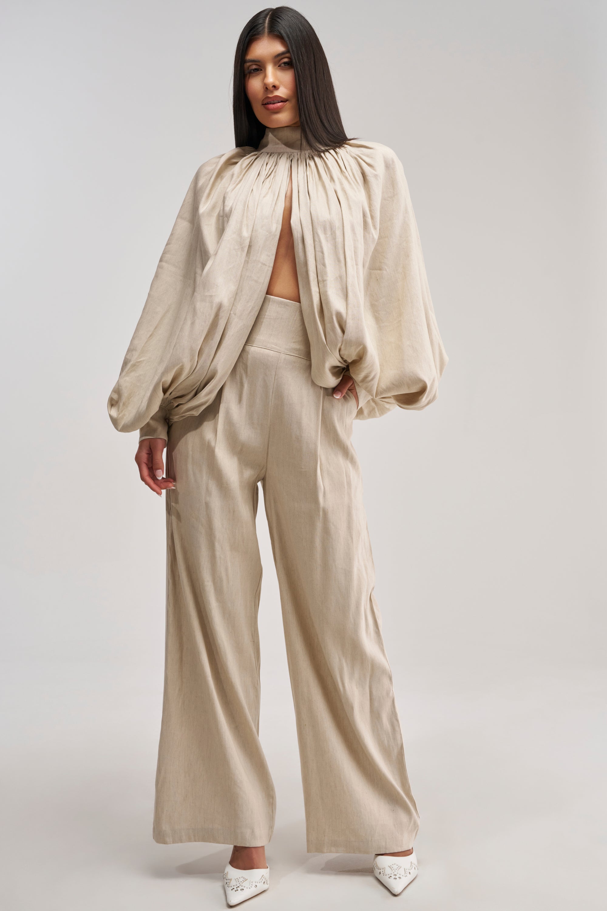 A woman stands in a beige, loose-fitting, long-sleeve top with gathered fabric at the neckline and a keyhole cutout. She pairs it with the MASTERPIECE WIDE LEG PALAZZO PANT IN LINEN for effortless summer date night styling. Her look is completed with white pointed shoes and straight hair.