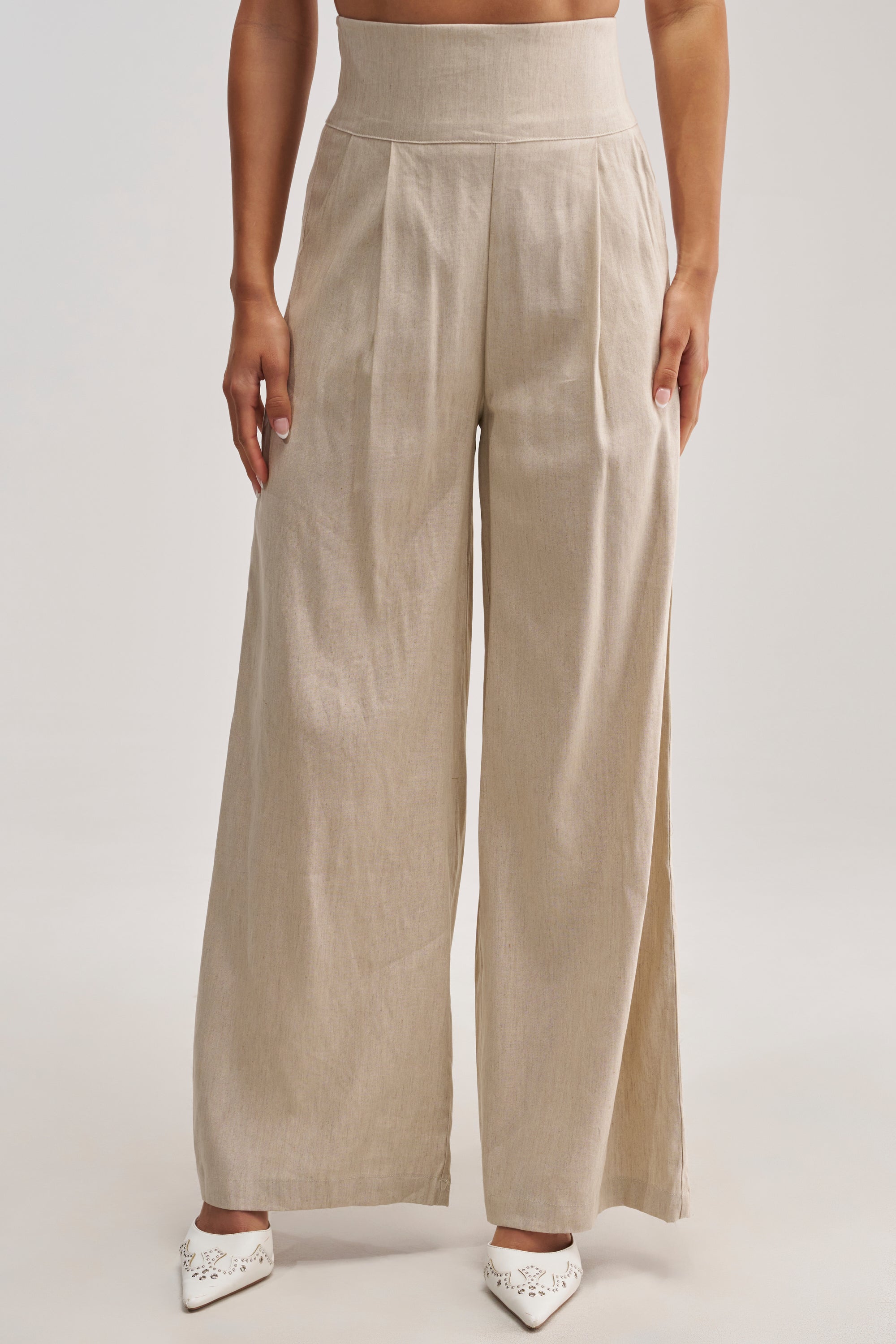 Dressed in the MASTERPIECE WIDE LEG PALAZZO PANT IN LINEN—high-rise, wide-leg beige pants featuring front pleats—and paired with white pointed-toe shoes, the individual stands against a plain background, embodying chic summer style perfect for an effortless date night.