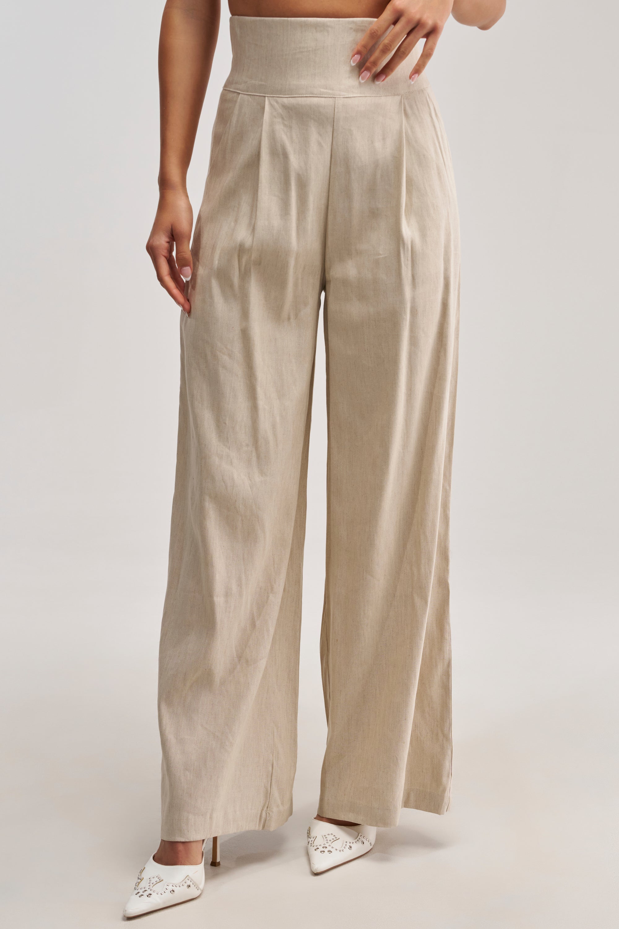 Styled for a summer date night, a person stands against a plain background with one hand on their waist, wearing the MASTERPIECE WIDE LEG PALAZZO PANT IN LINEN in beige, paired with white pointed-toe heels.