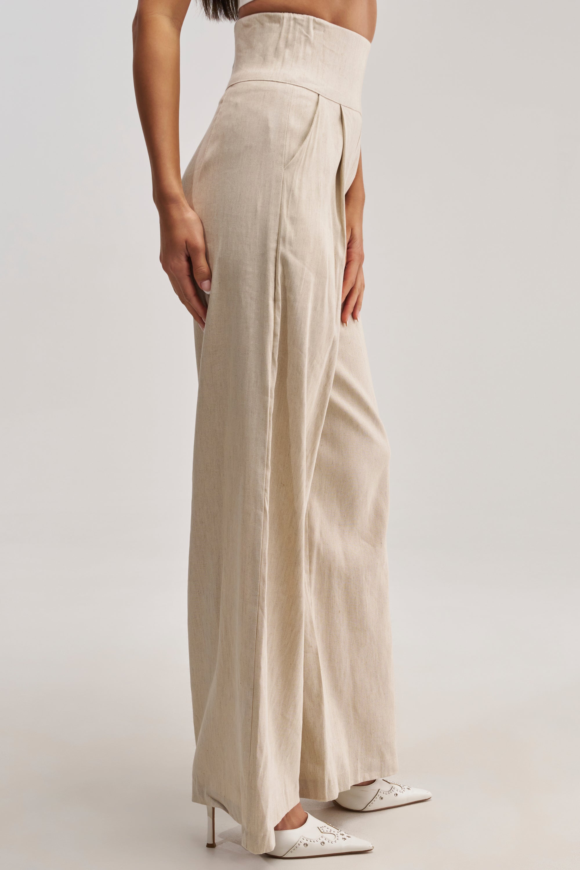 A person wearing the MASTERPIECE WIDE LEG PALAZZO PANT IN LINEN in a beige hue, paired with white pointed-toe heels, stands against a plain light background. The lower half of the body is highlighted, effortlessly showcasing summer date night style.