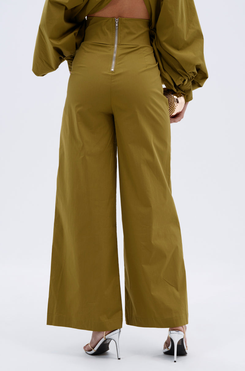 A person faces away wearing the GOLD COAST WIDE LEG PALAZZO PANT in olive green, featuring a visible back zipper. They pair it with silver high-heeled sandals and hold a small gold clutch. 4