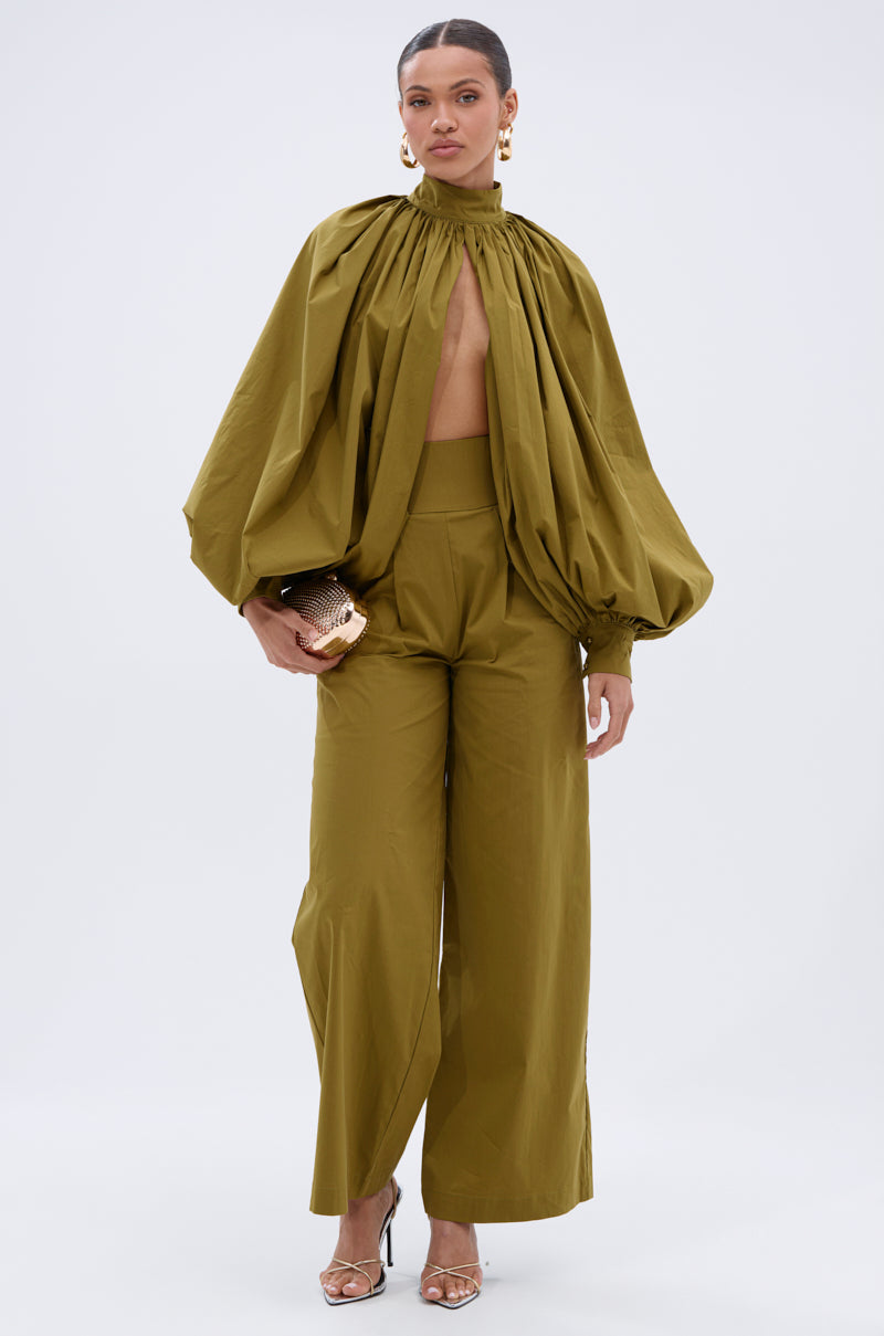 A woman stands against a plain light background in an olive green high-neck blouse, GOLD COAST WIDE LEG PALAZZO PANT, gold earrings, metallic heels, and a gold clutch. 6