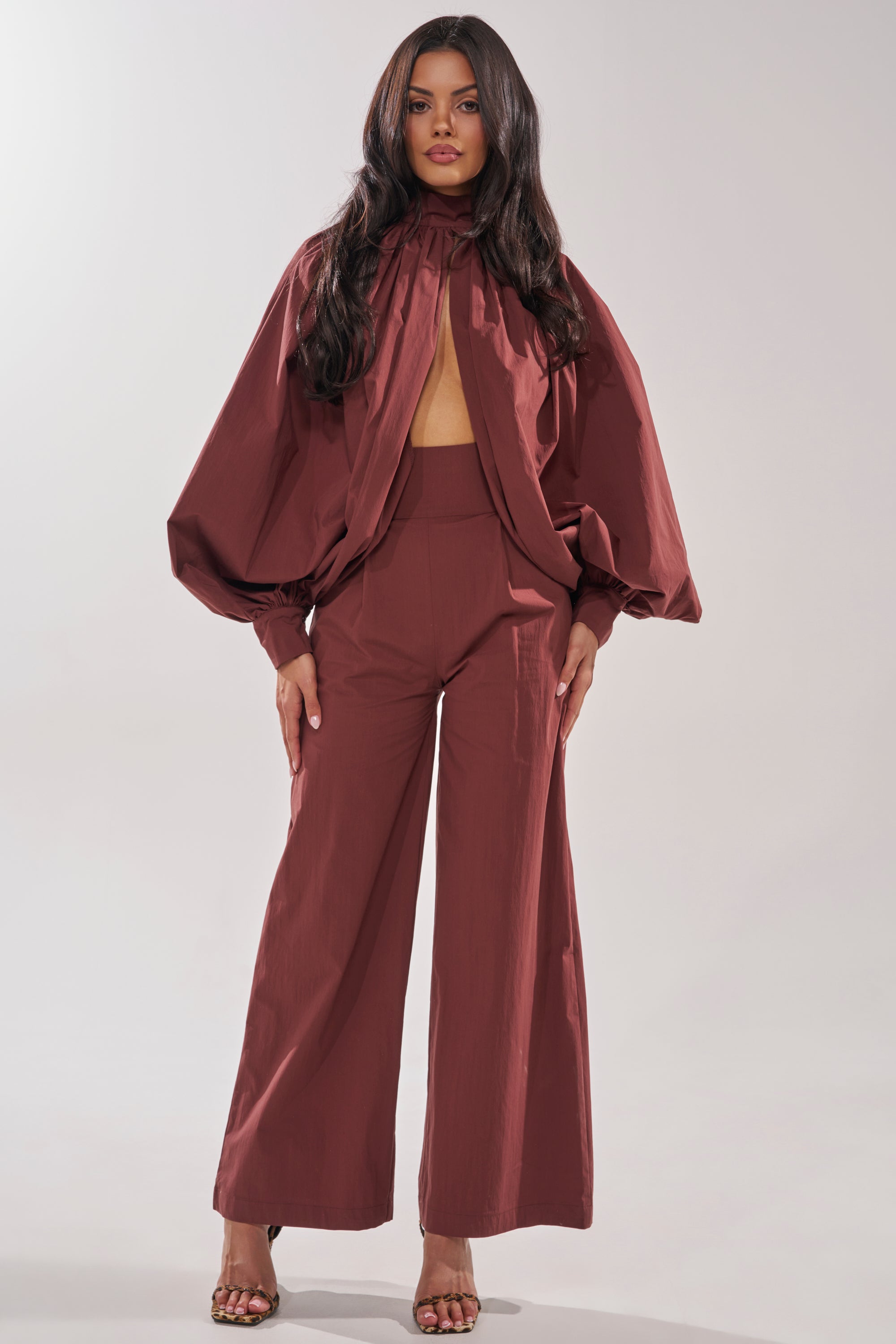 A woman faces forward in the GOLD COAST WIDE LEG PALAZZO PANT IN BROWN, a chic, loose-fitting style with high waist and wide legs. She pairs it with a matching voluminous sleeve top featuring a high neck and open front, finished with heeled sandals. 0